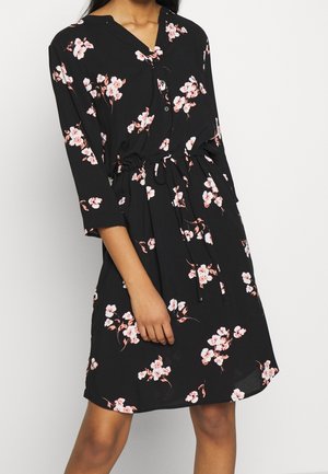 Černé šaty délky ke kolenům s růžovým a bílým květinovým potiskem, tříčtvrtečními rukávy, zapínáním na knoflíky vpředu a zavázaným páskem v pase na osobě.