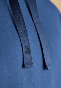 Gros plan sur des cordons de hoodie bleu foncé avec une texture en chevrons et un petit logo brodé sur un cordon, sur un fond de tissu bleu.