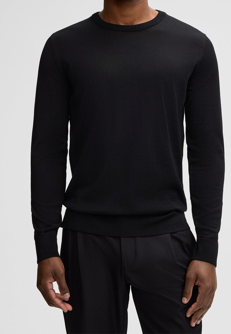 Homme portant un pull noir uni à manches longues avec col rond et un pantalon noir, debout devant un fond gris clair uni.