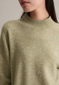 Pull en laine vert clair avec un col montant côtelé, une coupe décontractée et une texture douce. Présente une apparence légèrement mouchetée.