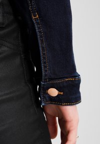 Mörkblå denimjacka med orange sömmar vid ärmsluten och en tryckknapp i kopparfärg, vilket lyfter fram dess rena design och texturerade tyg.