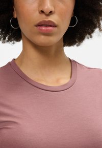 Lühikeste varrukatega, keha järgiva lõikega t-särk mauve värvi puuvillasegust. Omab ümmargust kaelaauku ja siledat tekstuuri. Pildil on nähtavad kõrvarõngad.