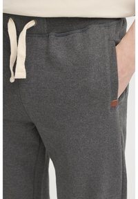 Grå fleece joggingbukser med snor i taljen, med sidelommer og et lille brunt logo-patch på venstre hofte. Blød tekstur.