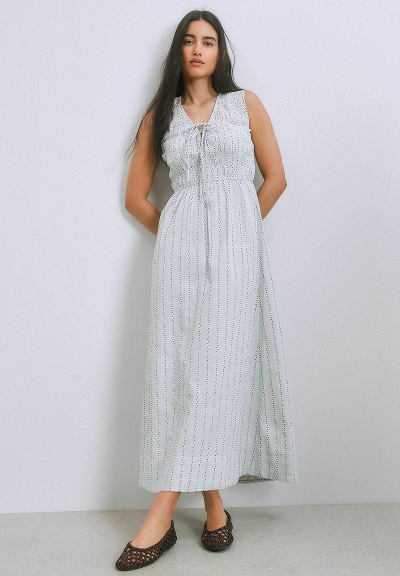 Nainen, joka pukeutuu hihattomaan valkoiseen maksimekkoon, jossa on pystysuoria pilkullisia raitoja, sekä mustiin punottuihin tasapohjaisiin kenkiin, seisoo yksinkertaista vaaleaa seinää vasten.