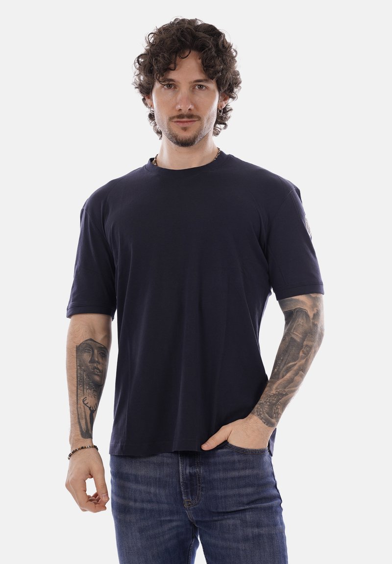 Uomo con capelli ricci e tatuaggi su entrambi gli avambracci indossa una maglietta blu scuro e jeans blu, in piedi con una mano in tasca.