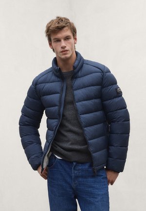 Young man wearing a navy blue quilted jacket, dark gray sweater, and blue jeans, standing against a plain light background.