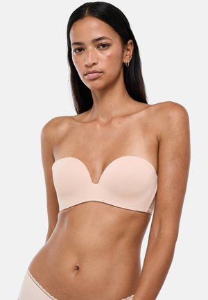 Femme aux longs cheveux foncés portant un soutien-gorge bandeau couleur chair et une lingerie assortie sur un fond clair uni.