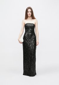 Juoda be petnešėlių suknelė su blizgučių dekoru, prigludusia silueto ir iki grindų ilgio. Derinama su nedidele balta audinio rankine.