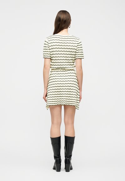 Γυναίκα στέκεται με την πλάτη προς τα εμάς, φορώντας ένα κρεμ φόρεμα με κοντά μανίκια και κυματιστές ρίγες, καθώς και μαύρες μπότες μέχρι το γόνατο, μπροστά σε λευκό φόντο.