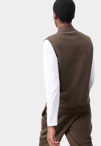 Brauner ärmelloser Pullover mit hohem Kragen, getragen über einem weißen Langarmshirt, mit einem sauberen Design und lässiger Passform.