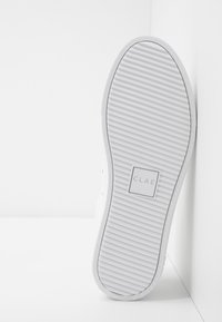 Semelle blanche de sneaker avec des rainures horizontales, logo rectangulaire avec l'inscription « CLAE » centré, positionné verticalement contre un mur d’angle blanc.