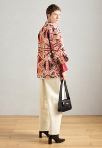 Színes, mintás, oversize dzseki geometrikus mintákkal, piros pólóval és krémszínű, bő szárú nadrággal párosítva. Kiegészítője egy fekete kézitáska.