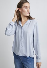 Blusa in poliestere azzurro chiaro con colletto, maniche lunghe e una vestibilità leggermente ampia. Chiusura semplice con bottoni, texture liscia e design minimale.