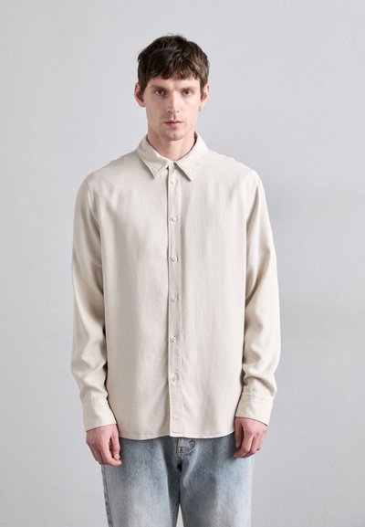 Chemise boutonnière beige clair avec un col classique, des manches longues et une coupe décontractée, fabriquée en tissu doux, portée avec un jean bleu clair.