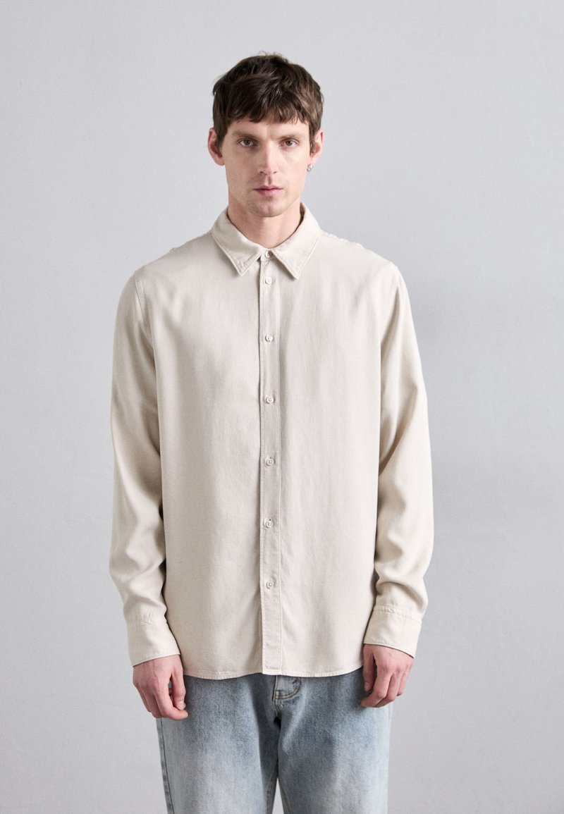 Cămașă button-up de un bej deschis, cu guler clasic, mâneci lungi și o croială relaxată, confecționată dintr-un material moale, purtată cu blugi albaștri deschiși.