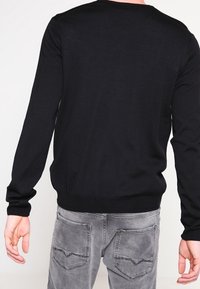 Maglione nero a maniche lunghe in morbido tessuto knit, con collo e orlo a coste, indossato con jeans in denim grigio chiaro.