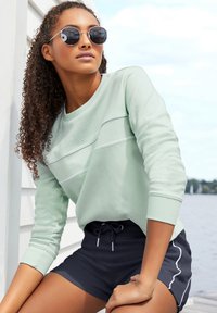 Muntgroene sweatshirt met horizontale paneel details en lange mouwen, gecombineerd met donkere shorts met witte bies. Ronde zonnebril maakt de look compleet.