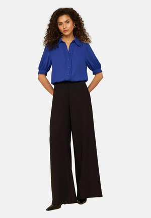 Vrouw met krullend haar, gekleed in een koningsblauw blouse met knopen en pofmouwen en wijde zwarte broek, staand met handen achter de rug.