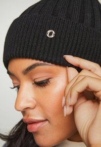 Gorro negro de punto con textura acanalada y acento de logotipo bordado en dorado. Vista de cerca del sombrero usado por una persona.