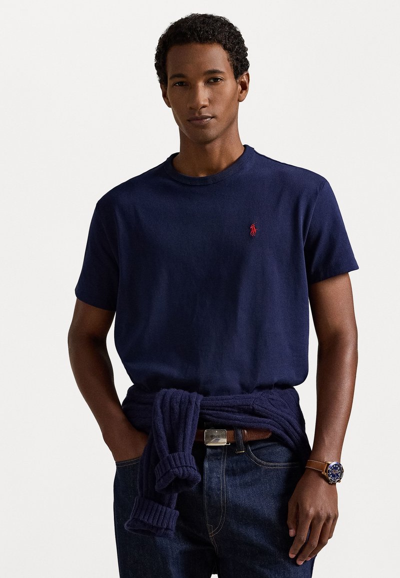 Navy kék, rövid ujjú póló piros logóval, pamut anyagból. Sötét farmernadrággal és derék köré kötött pulóverrel viselve. Barna öv és óra látható.