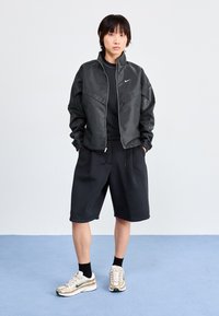 Svart vindjacka med dragkedja, med en Nike-logotyp. Matchad med svarta shorts och sneakers med guldaccent. Enkel, sportig design.