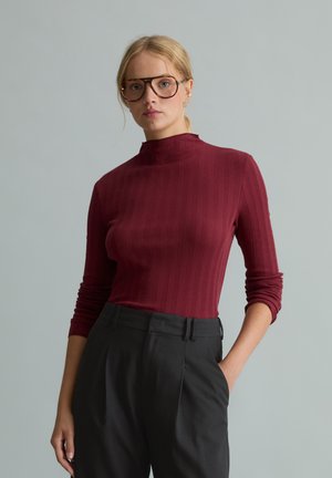 Vrouw met grote ronde bril, een bordeauxrode top met lange mouwen en zwarte hoge taille broek, staand tegen een effen grijze achtergrond.