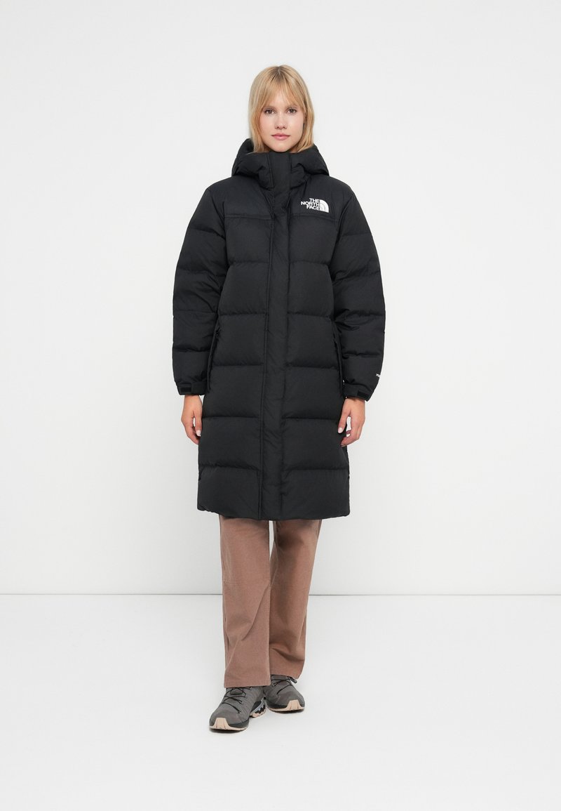 Crna puf jakna s kvadratnim uzorkom, visokim ovratnikom i logotipom The North Face na prsima. Uparena s smešnim hlačama i sivoj obući.