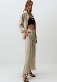 Blazer en lin beige sur un crop top noir, associé à un pantalon large beige et des sandales à talons noires. Texture lisse, coupe décontractée.