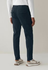 Celana kargo navy dengan banyak saku, desain menyempit, dan sedikit elastis. Dipadukan dengan sepatu sneaker abu-abu muda dan tan serta kaos lengan panjang putih.