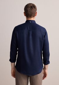 Camicia in lino blu navy a maniche lunghe, taglio morbido, orlo arrotondato e dettaglio con bottoni sul retro del collo, abbinata a pantaloni beige.