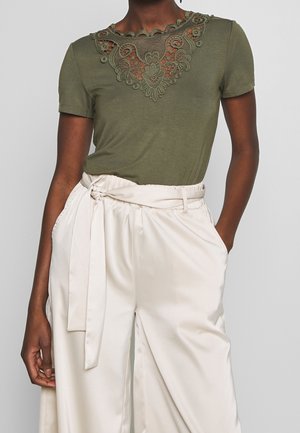 Kvinde iført olivengrøn kortærmet top med blomsterblondeudskæring ved halsen og højtaljede elfenbensfarvede satinbukser med bundet bælte.