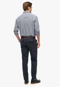 Uomo che indossa una camicia a maniche lunghe a quadri, pantaloni scuri con tasche posteriori, cintura marrone e scarpe nere, in piedi di spalle su sfondo bianco.