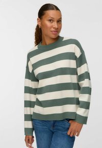 Donna con pelle marrone chiaro e capelli ricci raccolti, indossa orecchini a cerchio dorati, un maglione a righe larghe verde e bianco, e jeans blu.