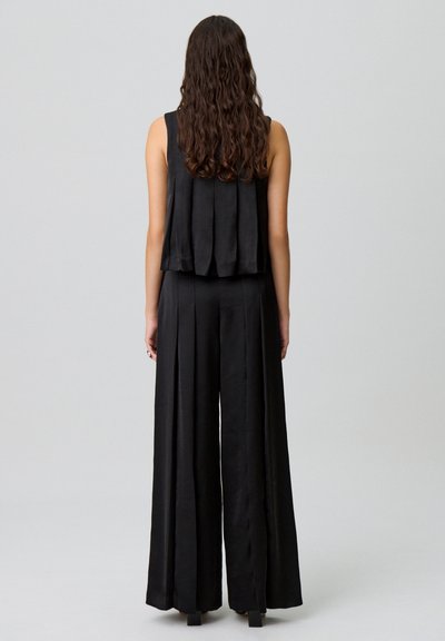 Mono de tela negra con diseño de pierna ancha, que presenta una parte superior plisada en la espalda y una textura suave. Su acabado brillante añade una elegancia sutil.