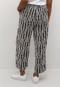 Personne portant un pantalon large à rayures ondulées noires et beiges, les mains dans les poches et des baskets blanches, se tenant devant un fond uni.