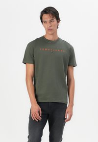 Jauns vīrietis ar olīvzaļu "Tommy Jeans" T-kreklu un tumši pelēkām biksēm, stāv uz viendabīga balta fona.