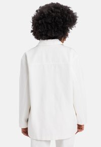 Veste en denim blanche avec une coupe décontractée, un col pointu et des manches longues. Le dos est uni avec un subtil détail de couture. Texture douce.