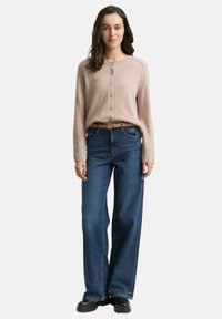 Lichtbruin langemouwen cardigan met knopen, gecombineerd met wijde donkere blauwe jeans en een tan riem. Glad textuur en casual pasvorm.