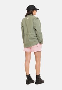 Femme portant une veste vert olive, un short rose, des bottes de combat noires et une casquette noire, debout les mains dans les poches, de dos.