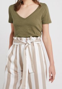 Hnědá v-neck tričko v olivové barvě spárované s vysoce pasovými kalhotami s bílými a béžovými pruhy a opaskem zavázaným na mašličku. Hladká textura.