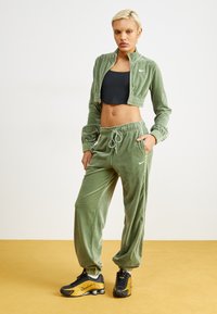 Chándal de terciopelo verde con chaqueta corta con cremallera y pantalones con cintura elástica. Presenta el logo de Nike, top corto negro y zapatillas deportivas negras y doradas.