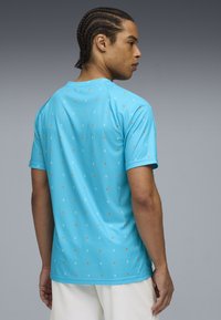 Turquāza īsajām piedurknēm krekls ar rakstainu dizainu, kurā ir mazi balti un oranži kvadrāti. Izgatavots no gludas, vieglas auduma.