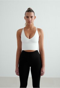 Hvid ribbet crop top med dyb V-udskæring og tynde stropper, parret med højtaljede sorte leggings. Glat tekstur, minimalistisk design.