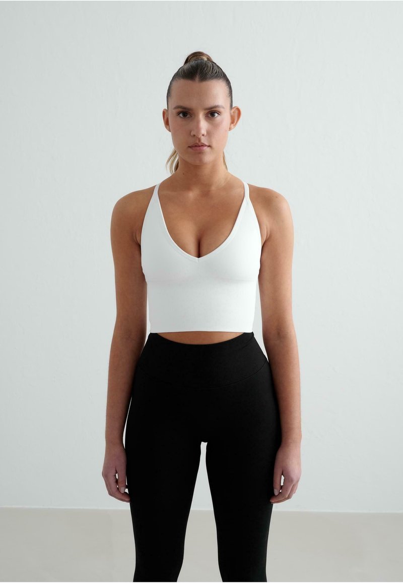 Hvid ribbet crop top med dyb V-udskæring og tynde stropper, parret med højtaljede sorte leggings. Glat tekstur, minimalistisk design.