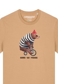 Beige Baumwoll-T-Shirt mit einem Bären in einem gestreiften Outfit, der Fahrrad fährt. Das Design beinhaltet eine rote Strickmütze und den Text "Tour de Franz."