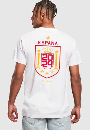 Valkoinen puuvillapaita, jossa on suuri punainen ja keltainen kilpimuotoilu selässä, jossa lukee "ESPAÑA" ja vuodet "2024" ja "2022".