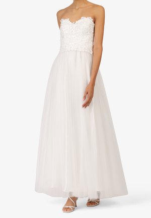 Schulterfreies weißes Kleid mit floraler Spitzen-Büste und fließendem Tüllrock, kombiniert mit weißen Riemchen-Pumps.
