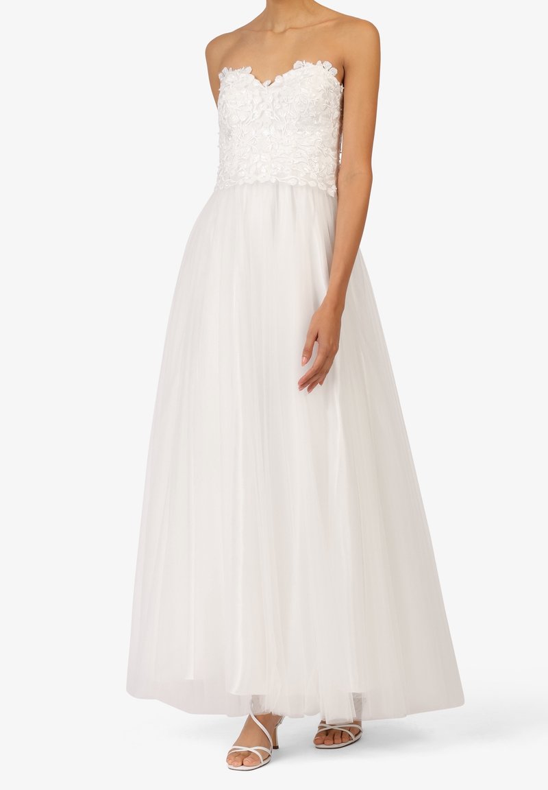 Robe blanche bustier en dentelle florale avec jupe en tulle fluide, portée avec des sandales blanches à talons et fines lanières.