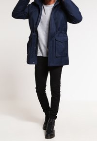 Parka de color azul marino con capucha, cierre delantero y dos grandes bolsillos frontales. Llevado sobre una camisa gris claro y combinado con jeans negros y botas.