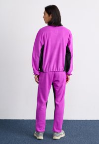 Fleece treningsdrakt i livlig lilla farge med svarte sidepaneler, avslappet passform og elastiske mansjetter. Merkbar logo ved kanten.